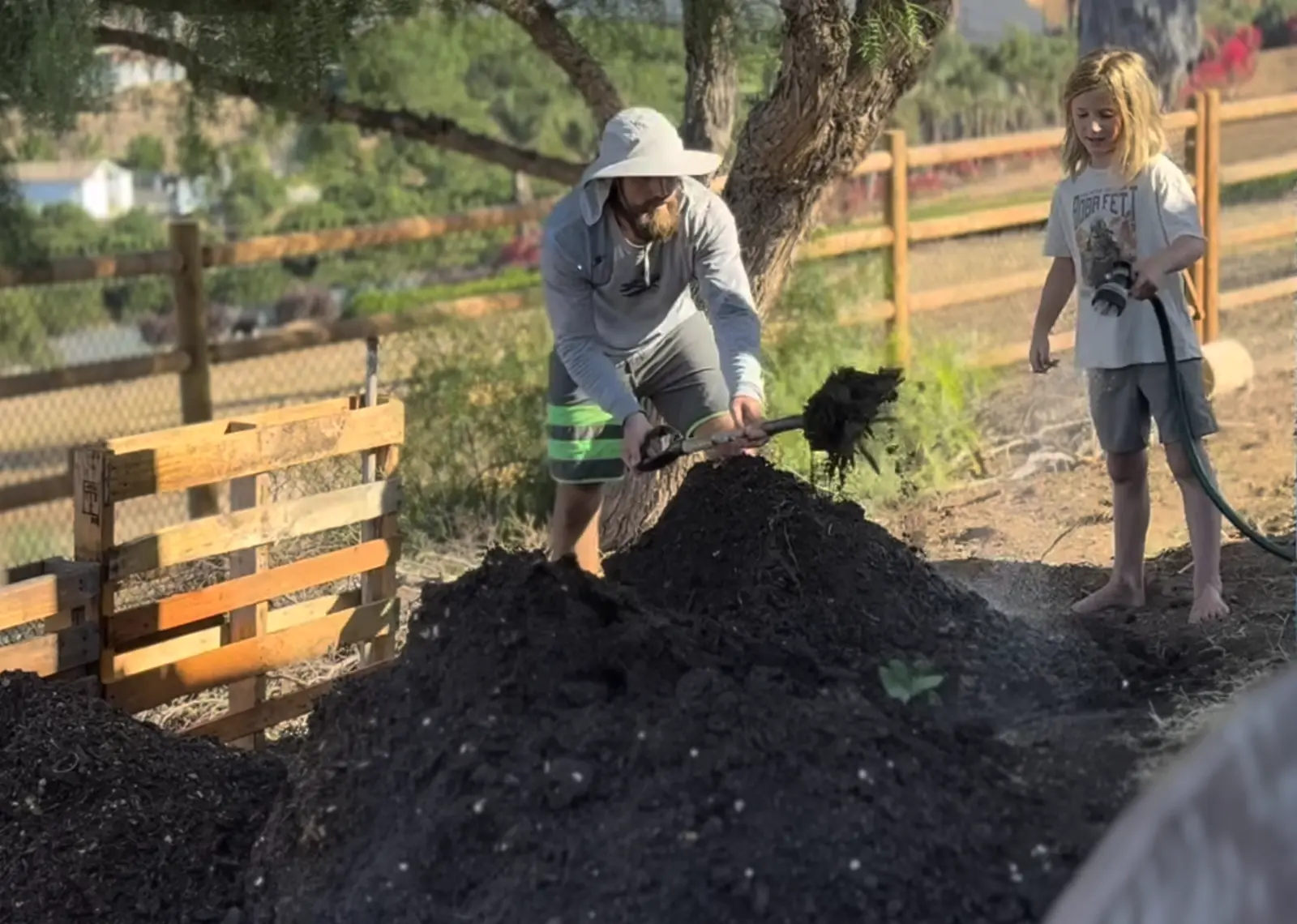 A father turning a large pile of dark, finished compost with a shovel while one of the children waters it down with a hose at Tradewinds Ranch