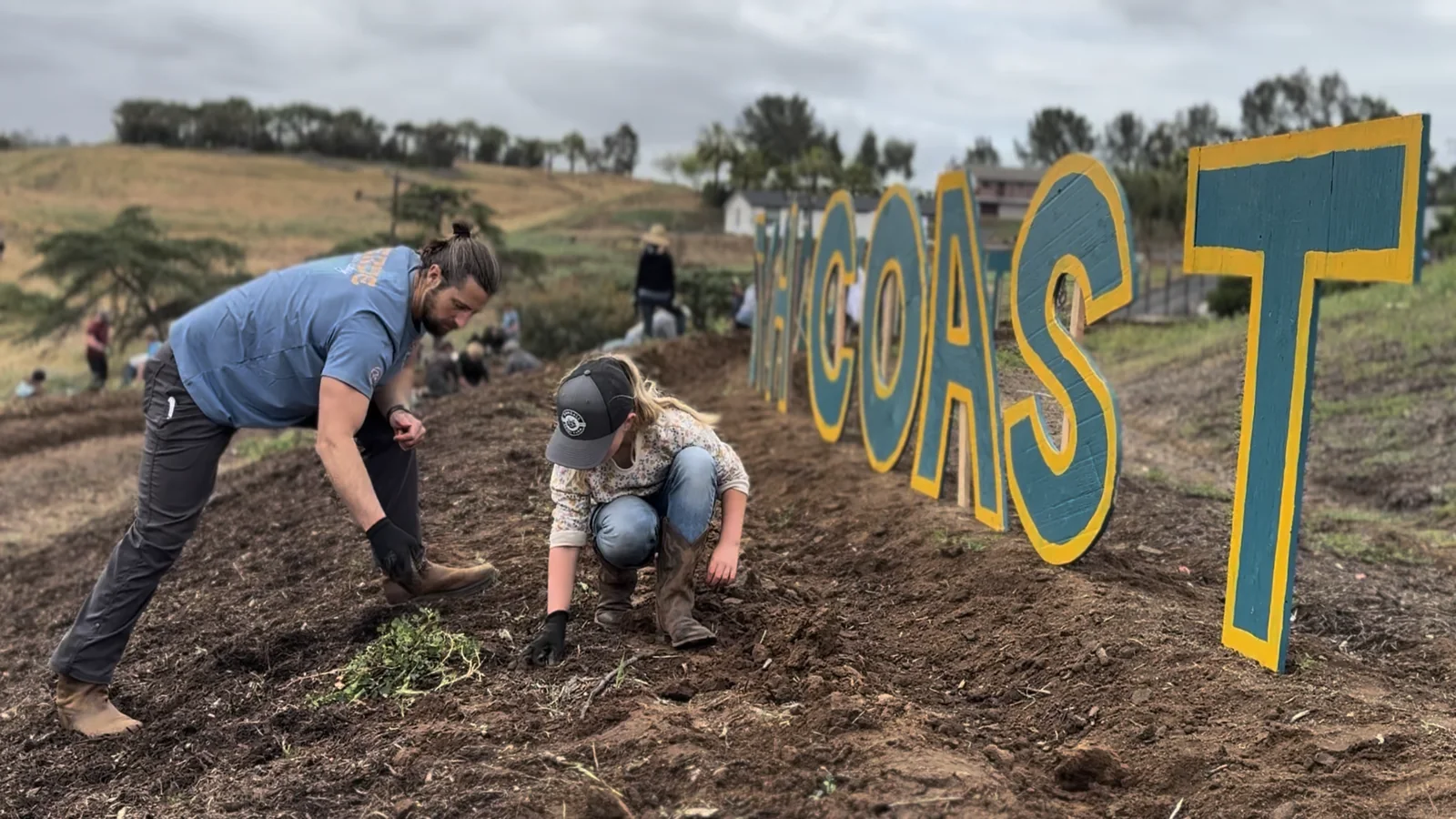 Neighbors working the soil together at a North County farm, with a hand-painted COAST sign standing in the field