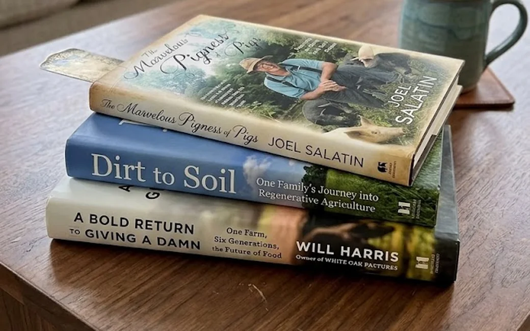 A stack of regenerative farming books, The Marvelous Pigness of Pigs, Dirt to Soil, and A Bold Return to Giving a Damn, on a wood coffee table