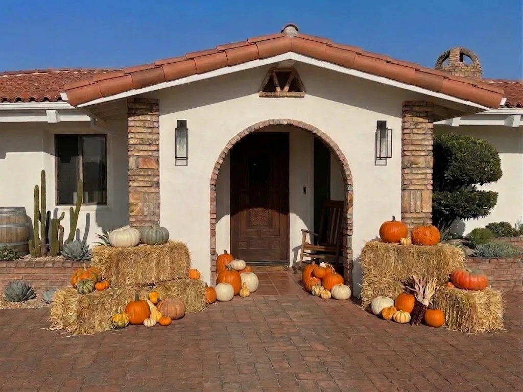 A finished fall porch display at a Spanish-style arched entry, hay bales and heirloom pumpkins flanking the brick walkway