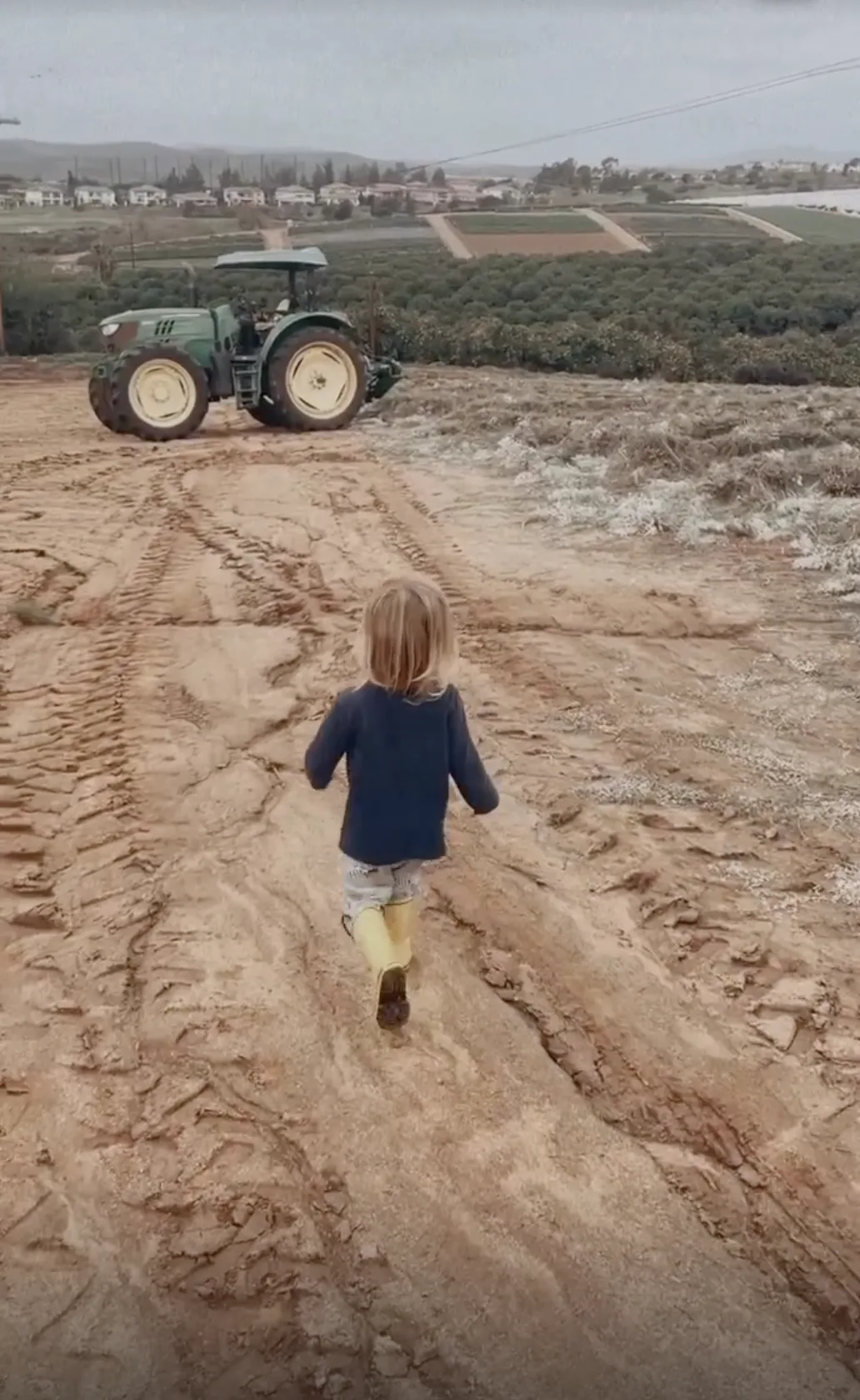 A toddler in rain boots walking down the wet dirt road toward the tractor