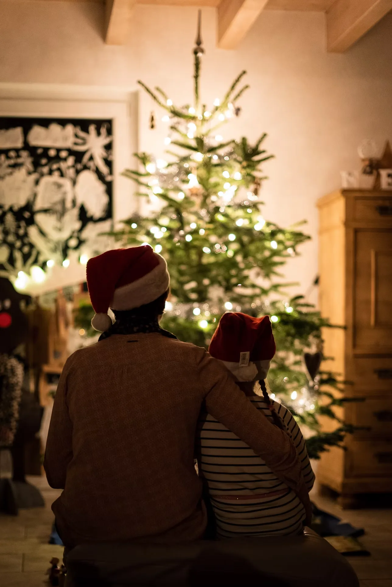 A parent and child in Santa hats sitting together looking at their lit Christmas tree