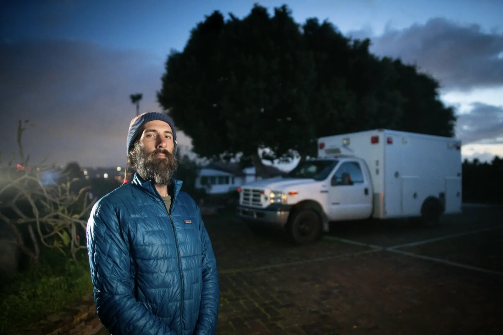 Sheena's brother in a blue jacket and beanie standing at dusk at Tradewinds Ranch, his converted ambulance camper parked under the big tree behind him and the valley lights below