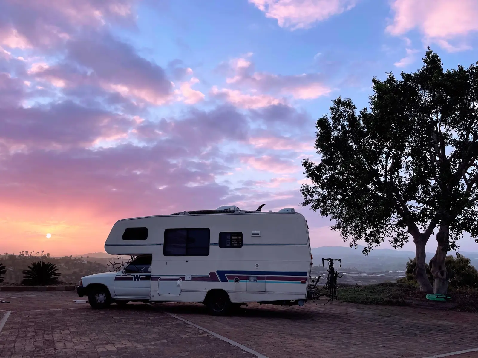 A vintage Toyota Winnebago motorhome parked on the brick pavers at Tradewinds Ranch at sunrise, a large tree beside it and the valley beyond under a soft pink and lavender sky