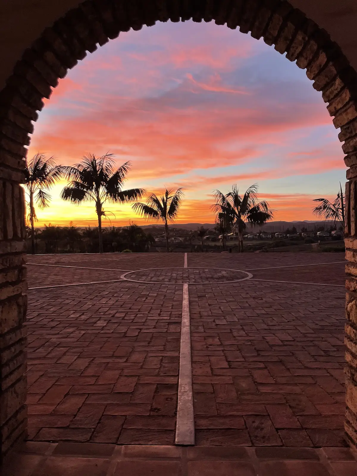 The view out through the brick entry arch at Tradewinds Ranch, a fiery pink and orange sunset over palms and the distant hills, the front courtyard pavers and the sundial line leading to the horizon